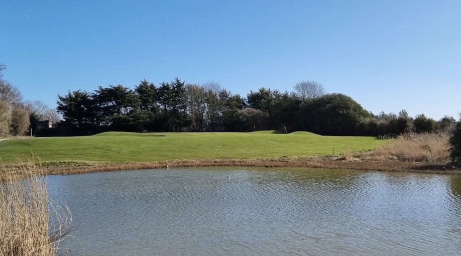 Great Salterns Municipal Golf Club, Hampshire, - View of golf course green from across large pond water feature, trees lining backdrop